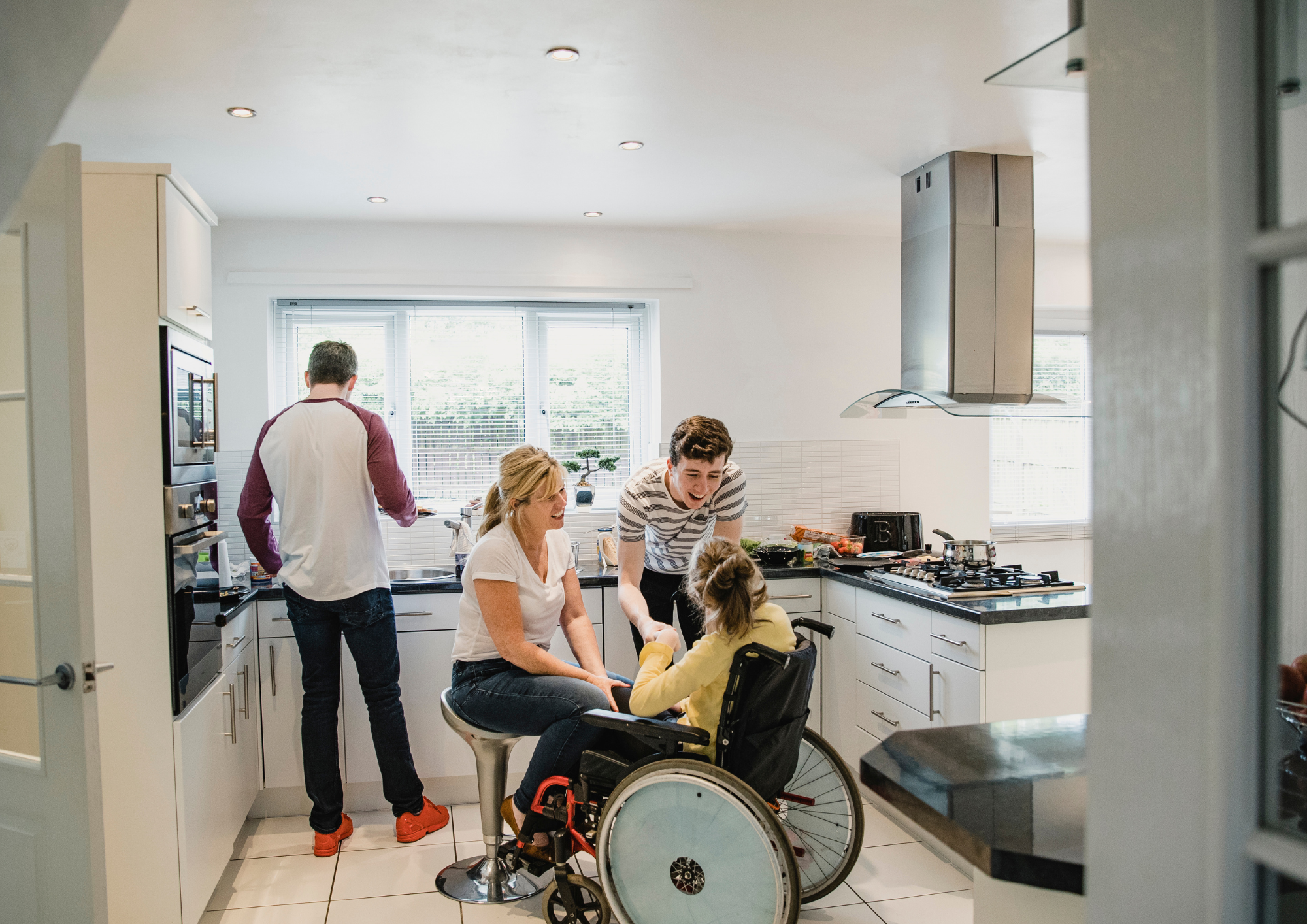 A man, woman, boy and girl are in the kitchen. The girl is in a wheelchair, the woman and boy chat to her as the man washes up at the kitchen sink.