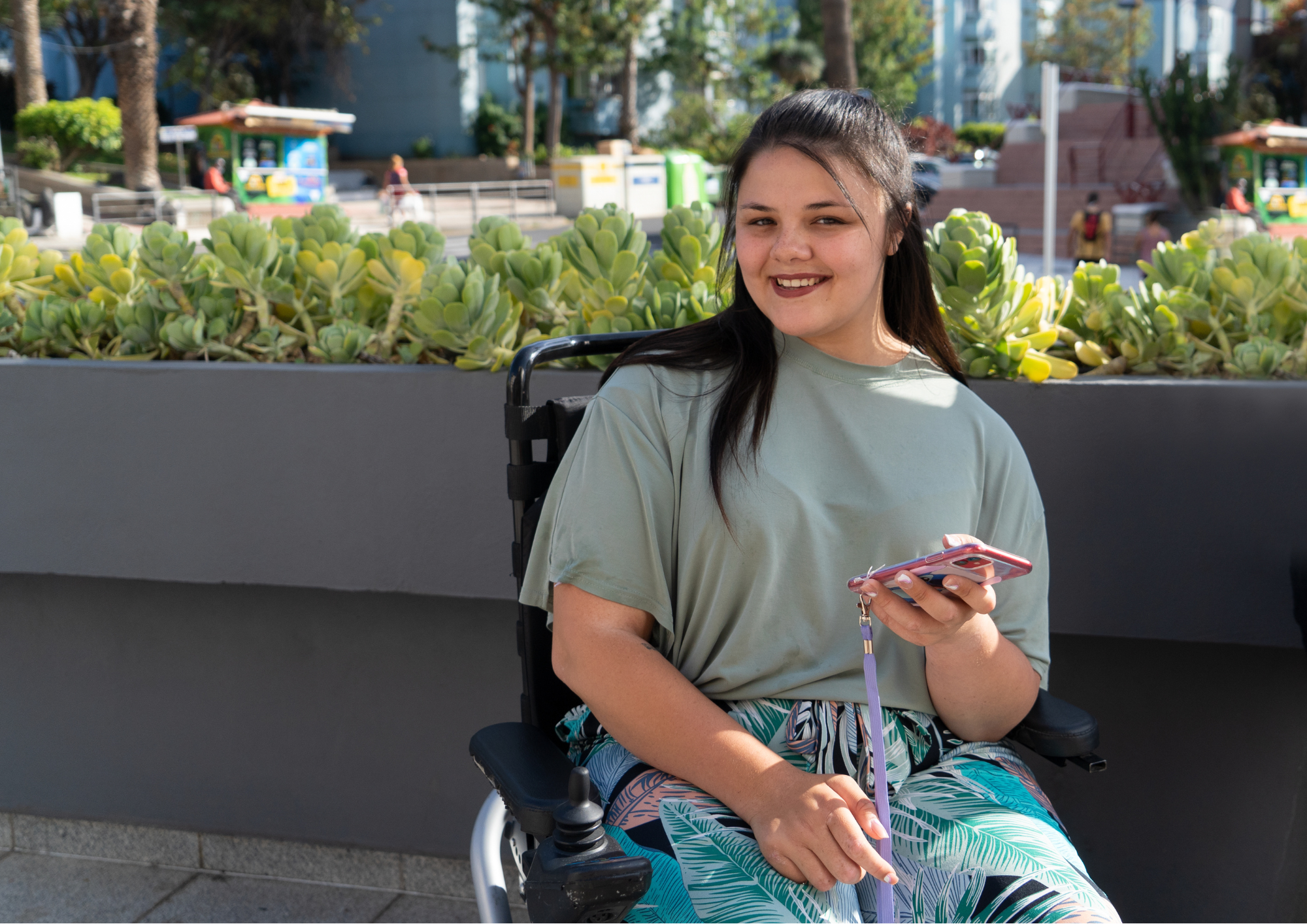 A young girl in a wheelchair smiles at the camera with a smartphone in hand.